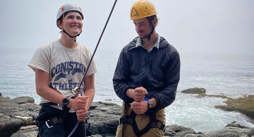 Two people wearing safety gear stand near a shore. One of them looks up as they belay an unseen climber. 